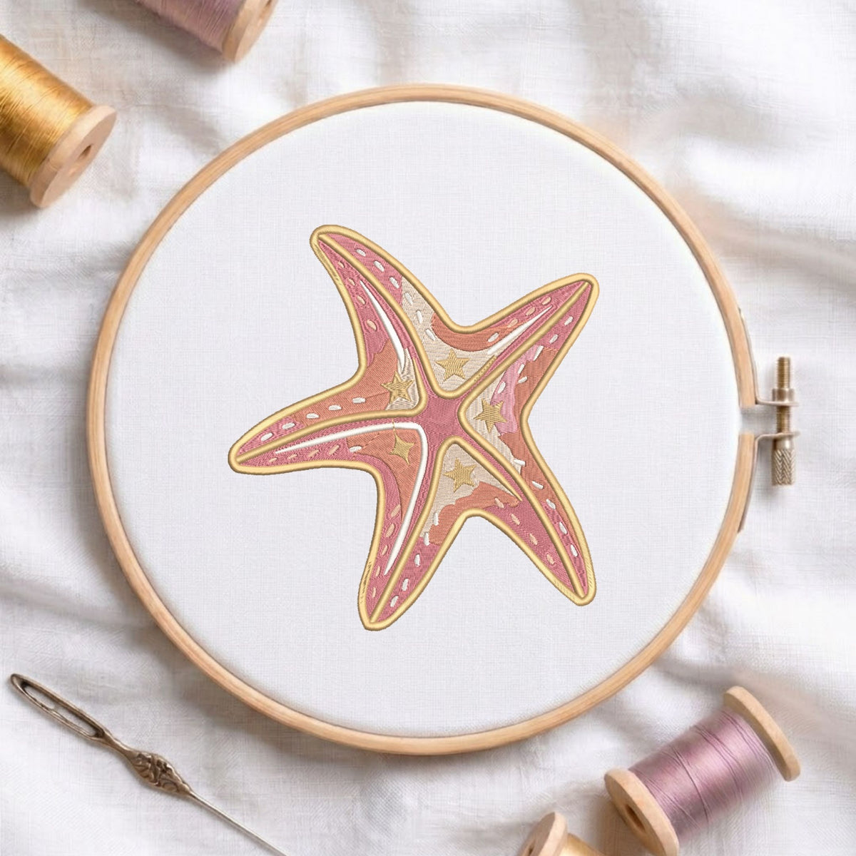 Pink and beige coastal starfish machine embroidery design displayed inside a round wooden hoop on white fabric alongside thread spools and a needle.