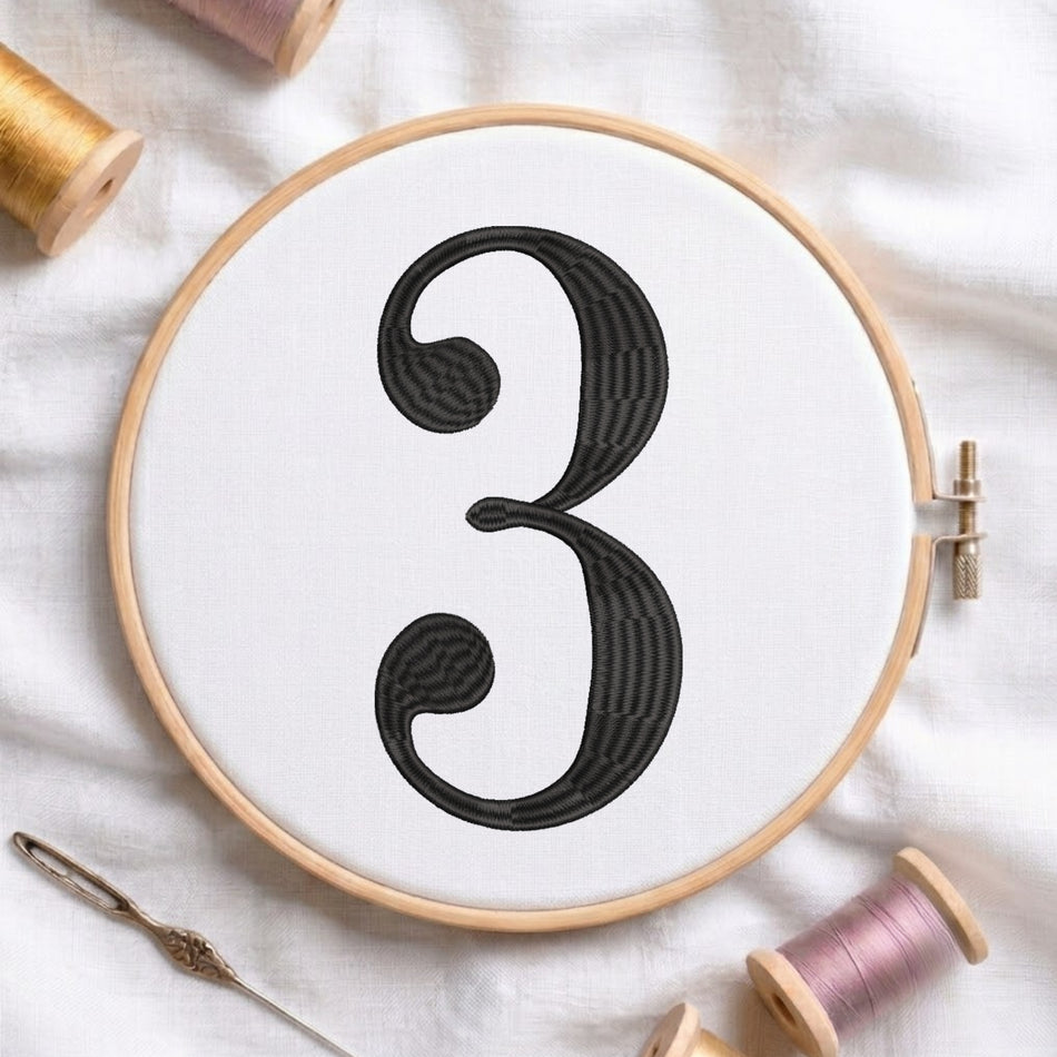 A wooden embroidery hoop holds white fabric with a large black stitched number 3. A needle and thread spools are arranged around it on a white fabric background.