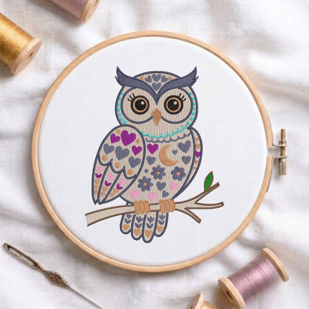 A circular embroidery hoop holding white fabric with a colorful embroidered owl design on a branch. Spools of thread and a needle are arranged around it on a white fabric background.