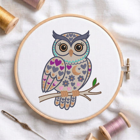 A circular embroidery hoop holding white fabric with a colorful embroidered owl design on a branch. Spools of thread and a needle are arranged around it on a white fabric background.