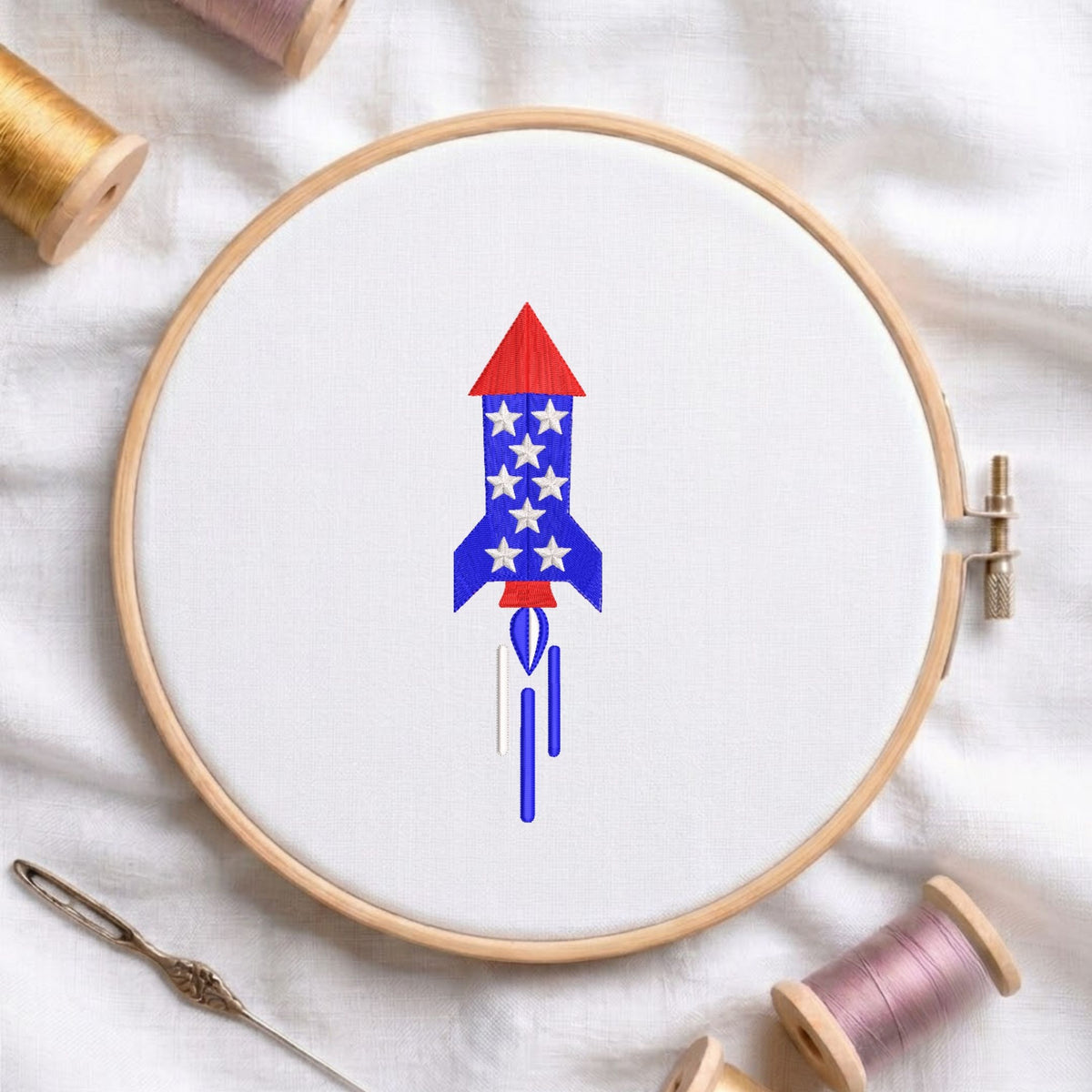 Red, white, and blue patriotic space rocket with star patterns launching with smoke trails machine embroidery design, displayed inside a round wooden hoop on white fabric with thread spools and a needle.