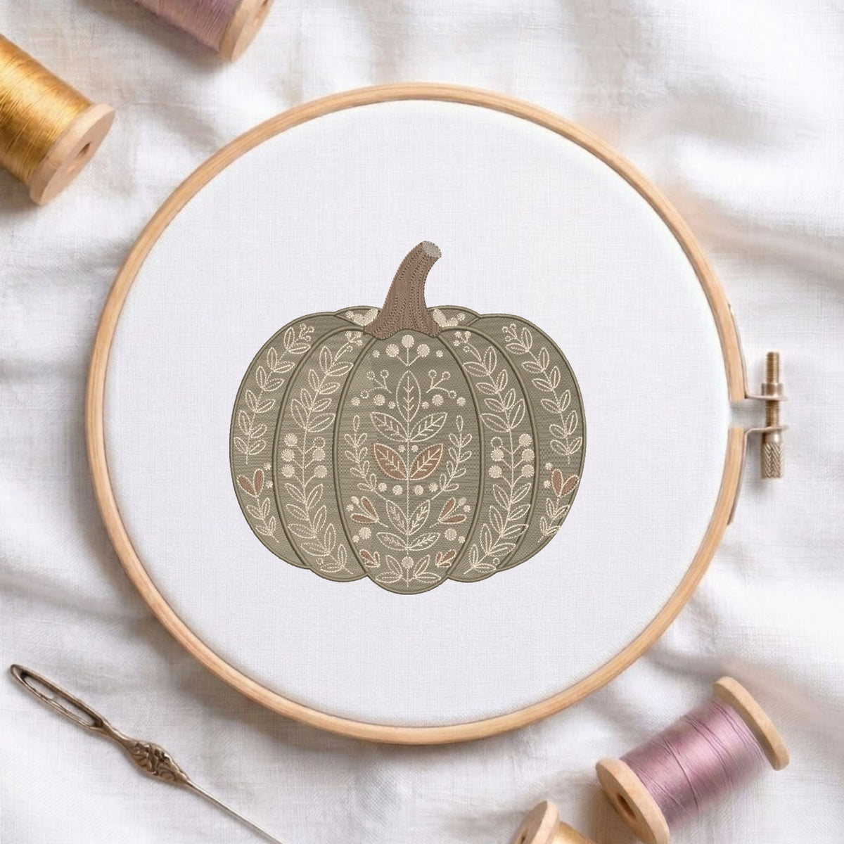 Botanical leaf-patterned pumpkin machine embroidery design displayed in a wooden hoop on white fabric with thread spools and a needle