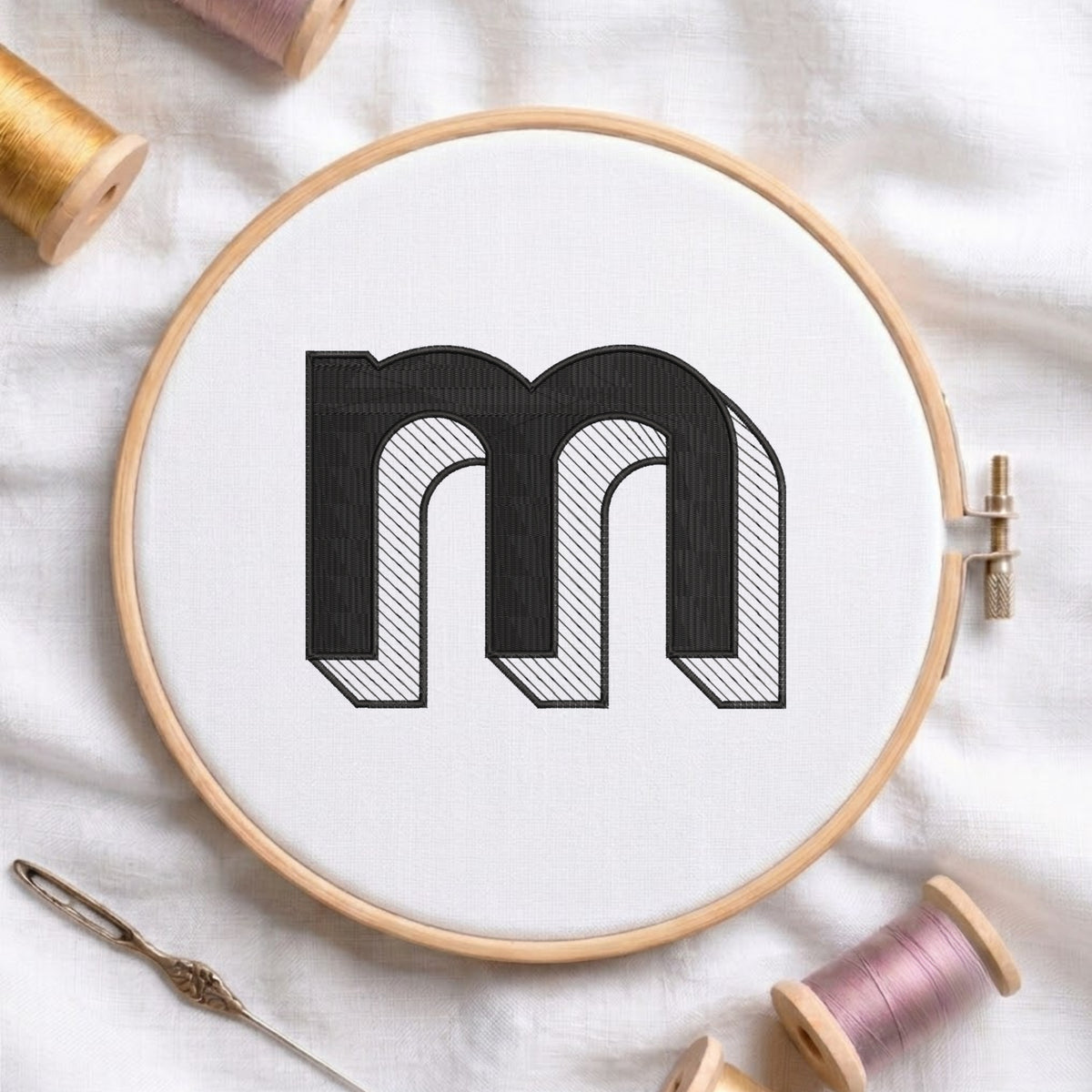 A wooden embroidery hoop holds white fabric with a large black embroidered letter 'm' outlined in black and filled with fine diagonal hatching. Spools of thread, a needle, and soft fabric are arranged around it in a styled flat-lay.