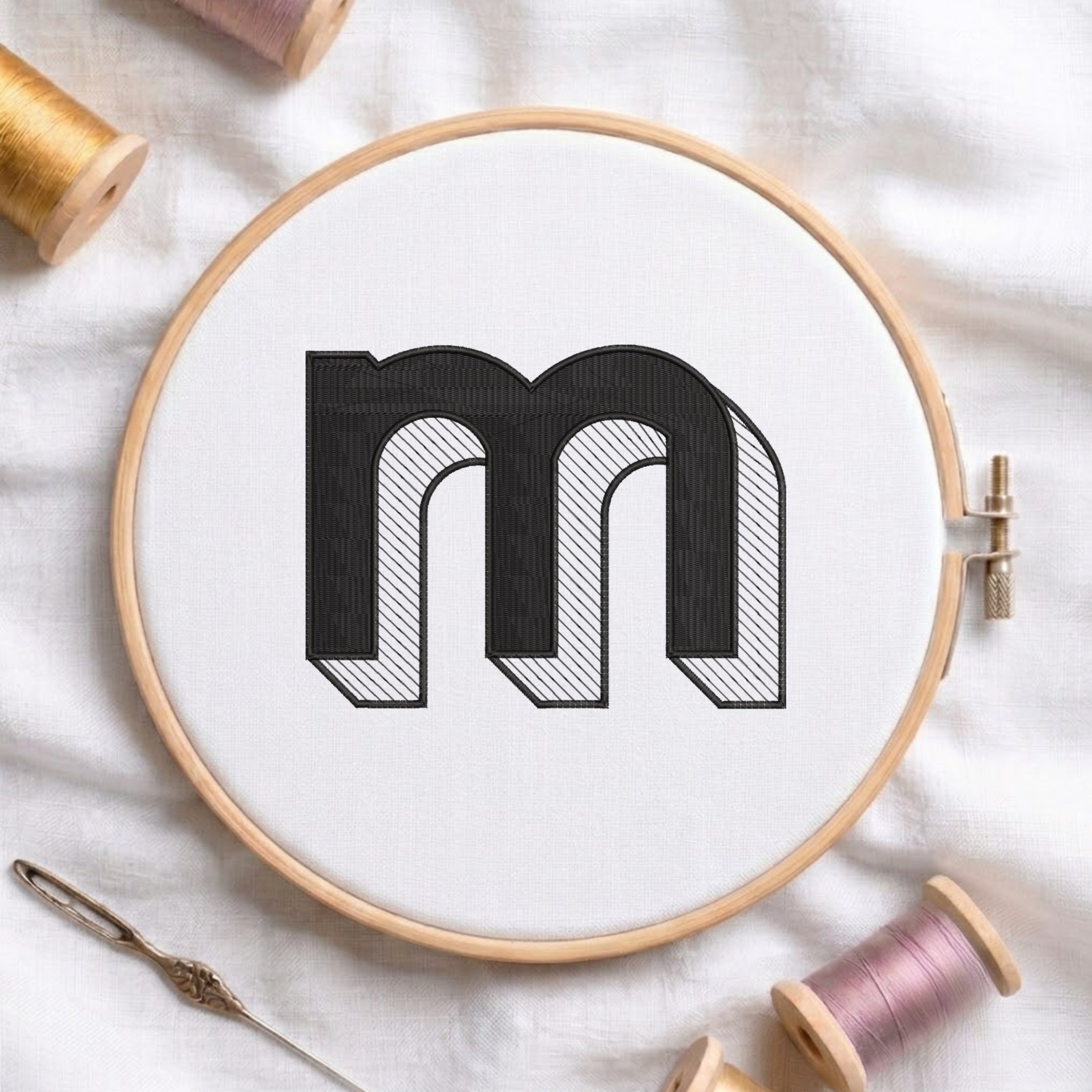 A wooden embroidery hoop holds white fabric with a large black embroidered letter 'm' outlined in black and filled with fine diagonal hatching. Spools of thread, a needle, and soft fabric are arranged around it in a styled flat-lay.