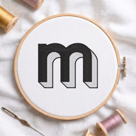 A wooden embroidery hoop holds white fabric with a large black embroidered letter 'm' outlined in black and filled with fine diagonal hatching. Spools of thread, a needle, and soft fabric are arranged around it in a styled flat-lay.