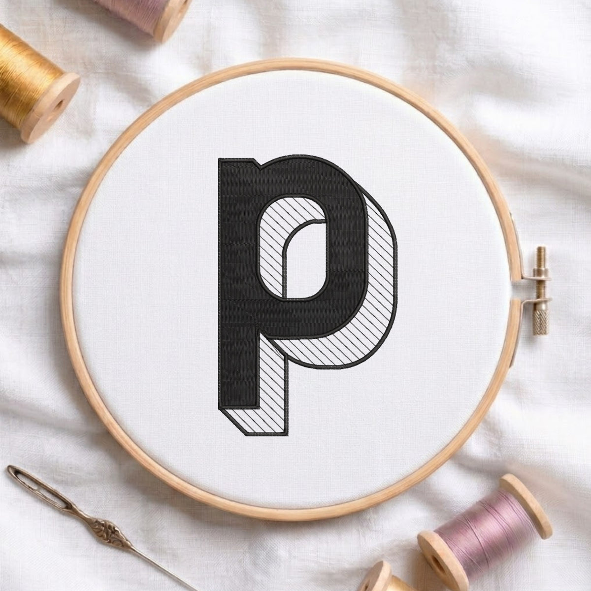 A wooden embroidery hoop holding white fabric with a large black letter 'p' stitched in the center. A needle lies at the lower left, and spools of thread are arranged around the hoop on a white fabric background.
