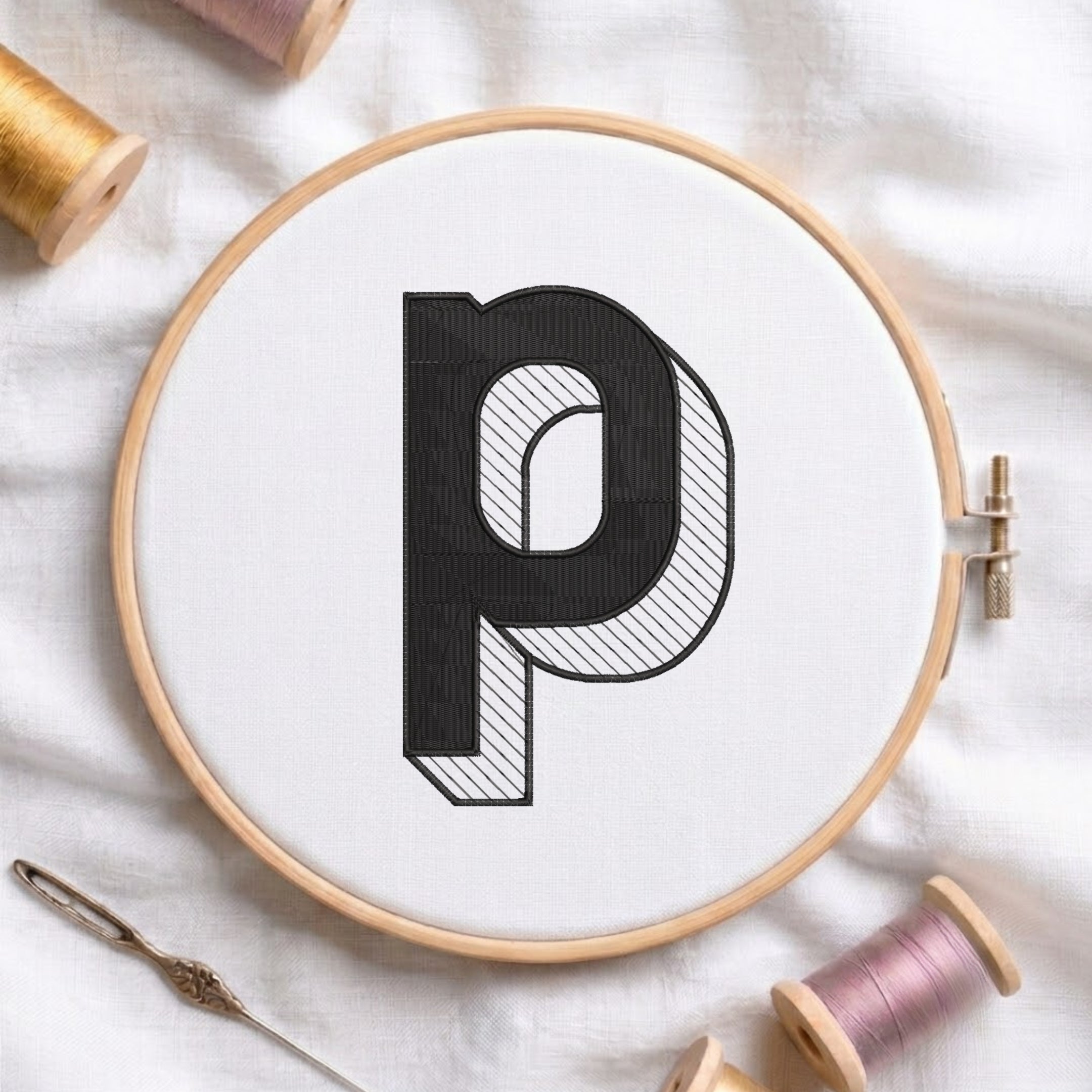 A wooden embroidery hoop holding white fabric with a large black letter 'p' stitched in the center. A needle lies at the lower left, and spools of thread are arranged around the hoop on a white fabric background.