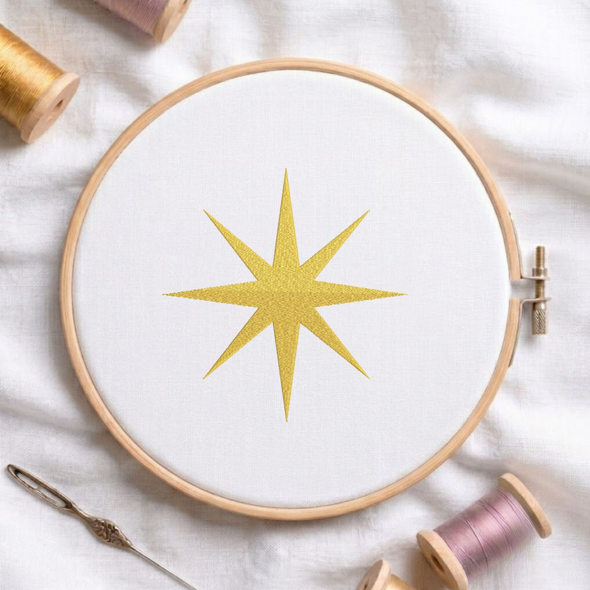 Golden swirling star machine embroidery design displayed in a wooden hoop on white fabric with thread spools and a needle