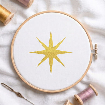 Golden swirling star machine embroidery design displayed in a wooden hoop on white fabric with thread spools and a needle