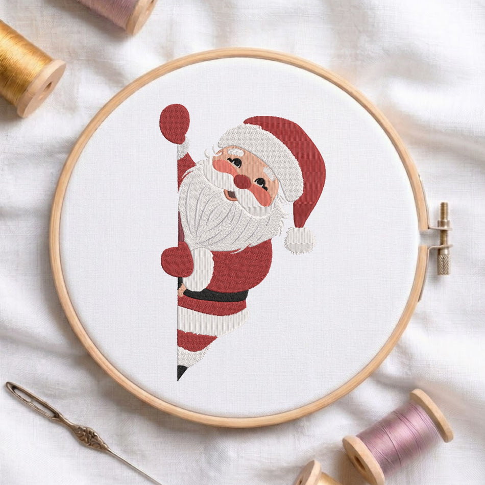 A white fabric stretched in a wooden embroidery hoop, featuring an embroidered cute Santa Claus peeking from the side with a red hat, white beard, and waving hand.