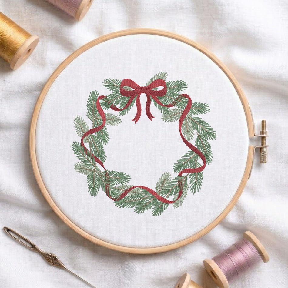 Christmas wreath embroidery design displayed in a round wooden hoop on a white linen fabric background with spools of thread and a needle.