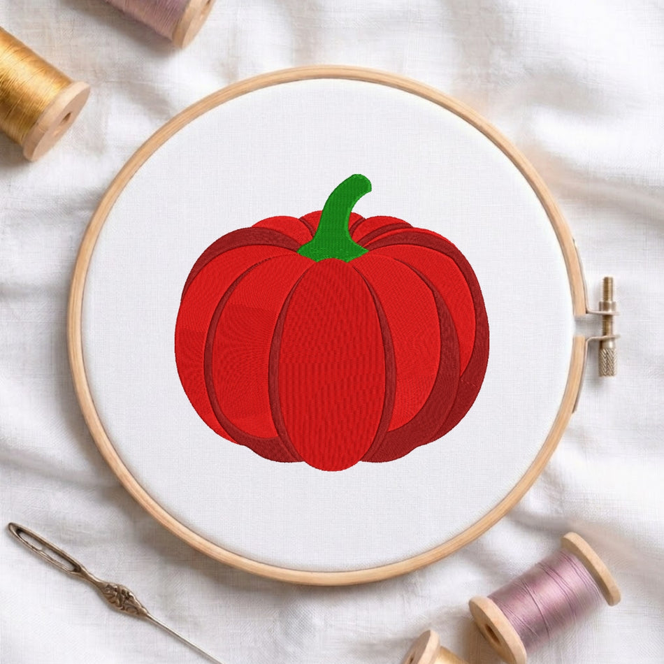 A circular embroidery hoop holding white fabric with a red pumpkin design and green stem, styled on a white cloth with thread spools and a needle around it.