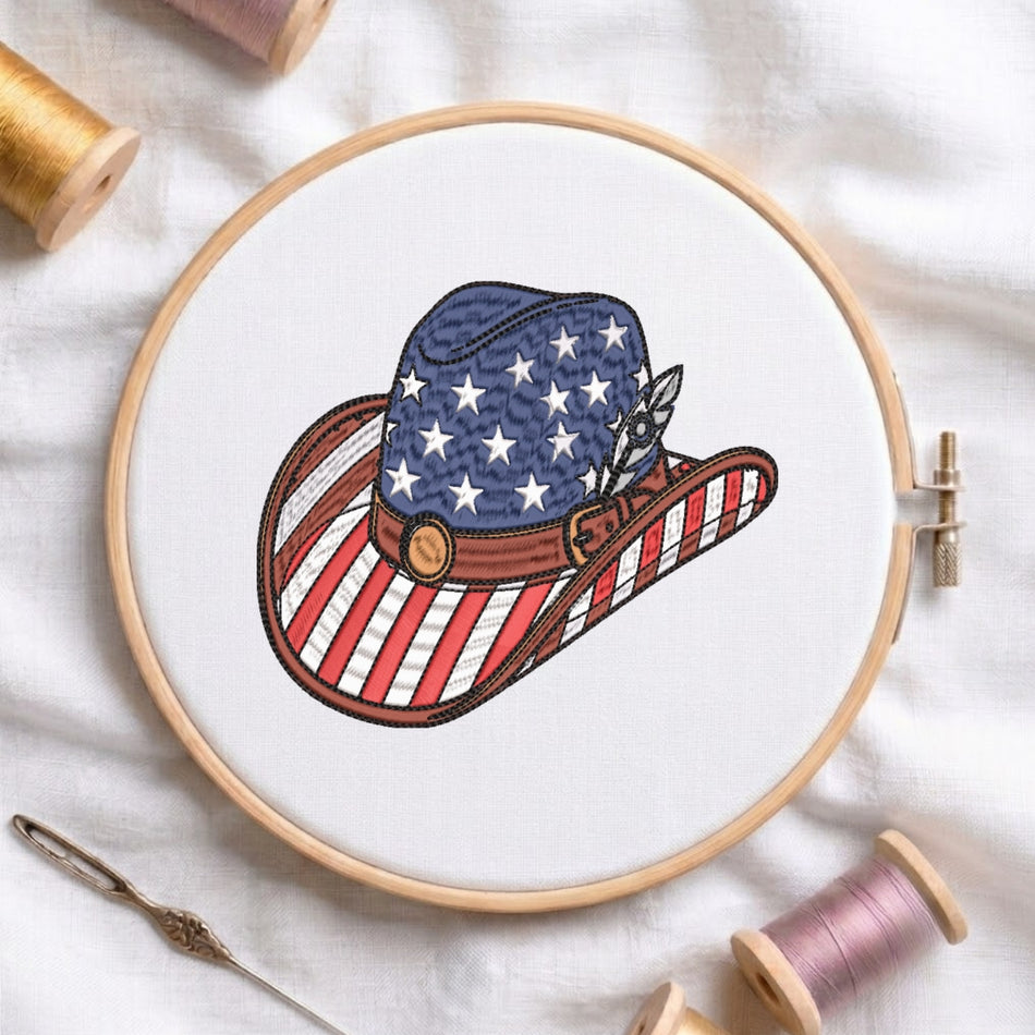 A circular embroidery hoop holding white fabric with a stitched cowboy hat design in American flag colors: blue with white stars on the crown, red and white stripes on the brim, and a brown band with a gold button. Spools of thread and a needle are arranged around it on a white fabric background.