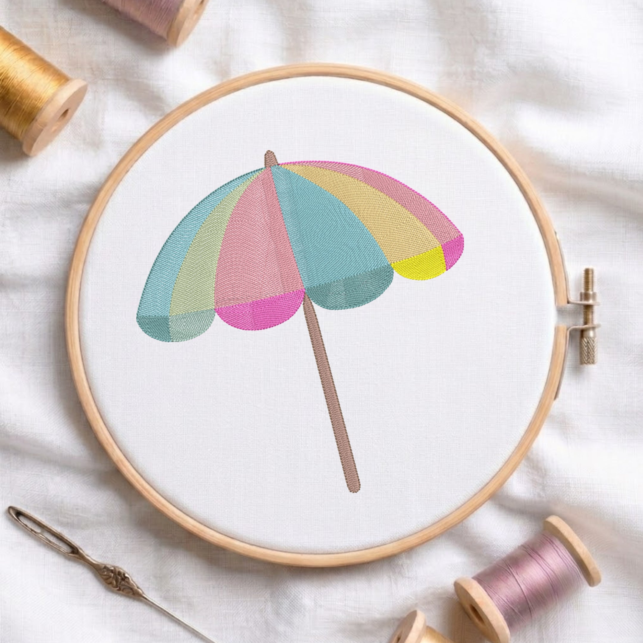 A pastel beach umbrella embroidery design held in a wooden embroidery hoop, surrounded by spools of thread and a needle on a white fabric background.