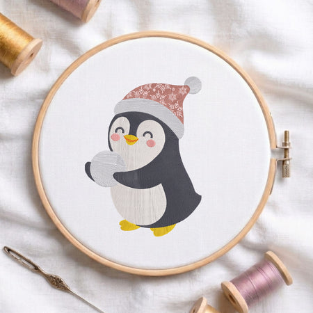 A circular embroidery hoop holding white fabric with a cartoon penguin wearing a patterned red Santa hat and yellow shoes. The hoop is styled on a white fabric background with spools of thread, a needle, and a wooden stitch comb nearby.