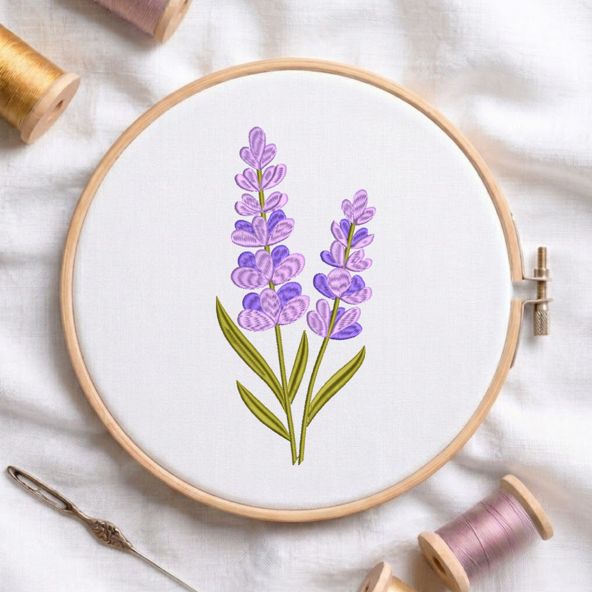 A wooden embroidery hoop holding white fabric with a lavender floral design in purple and green, surrounded by spools of thread and a needle on a white fabric background.