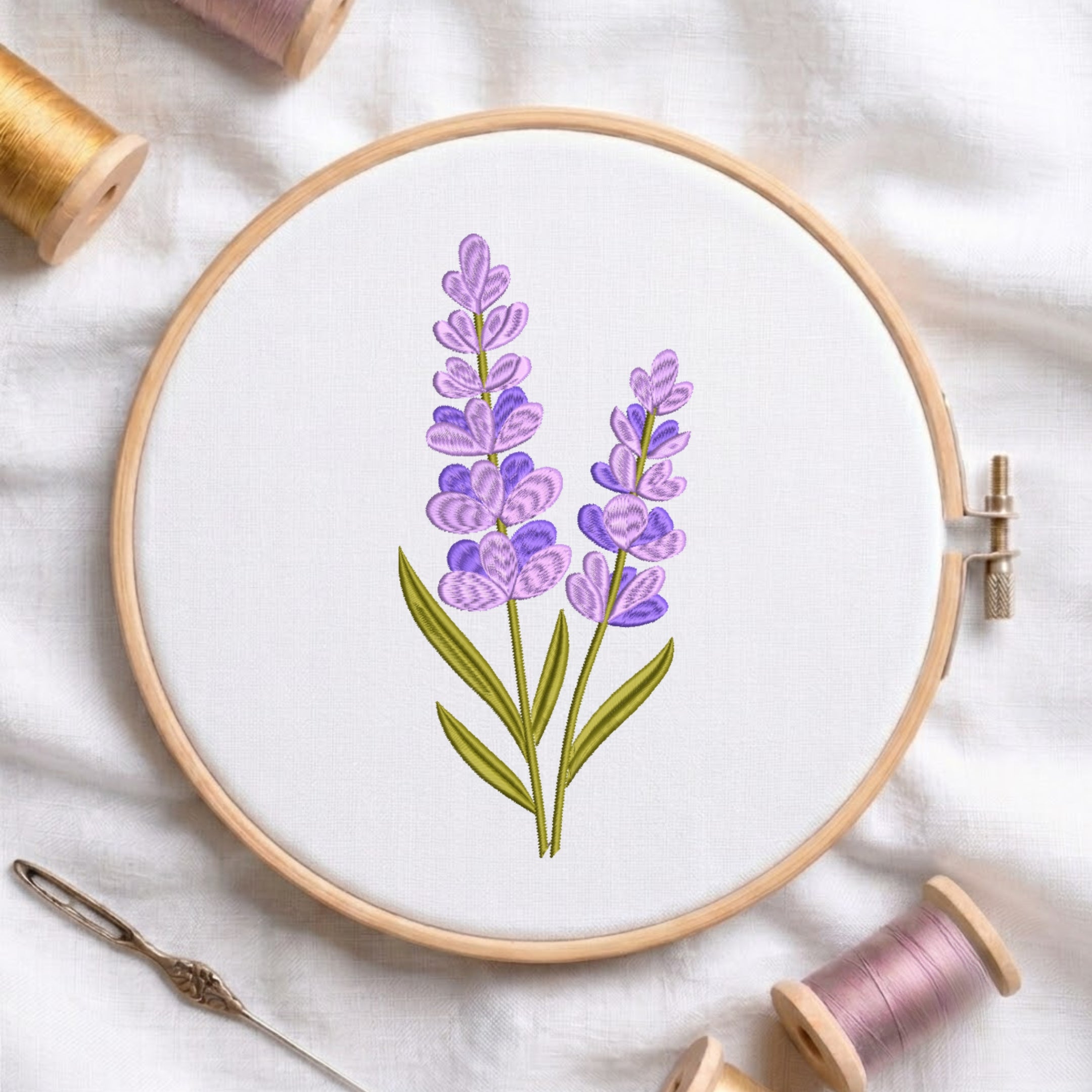 A wooden embroidery hoop holding white fabric with a lavender floral design in purple and green, surrounded by spools of thread and a needle on a white fabric background.