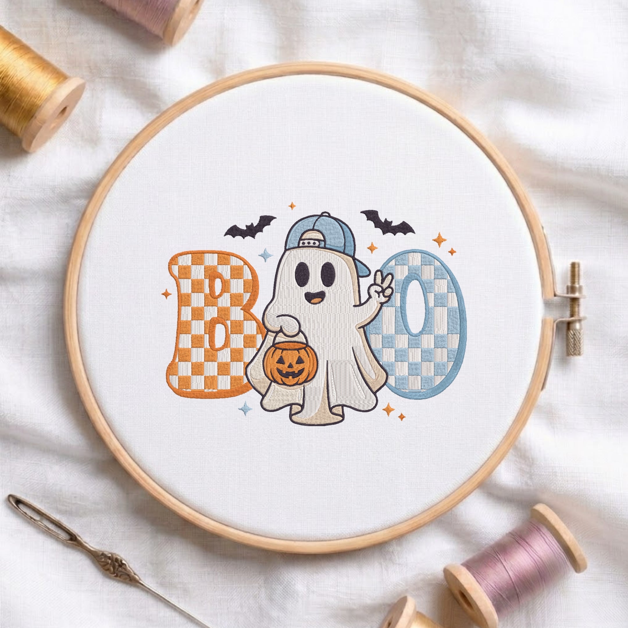 A circular embroidery hoop on white fabric displays a Halloween-themed 'BOO' design with a cartoon ghost holding a pumpkin. Spools of thread and a needle are arranged around the hoop.