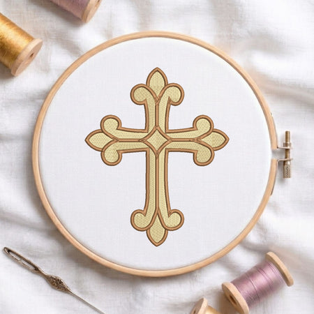 A wooden embroidery hoop holding white fabric with a decorative cross design stitched in beige and brown thread. A needle and spools of thread are arranged around it on a white fabric background.