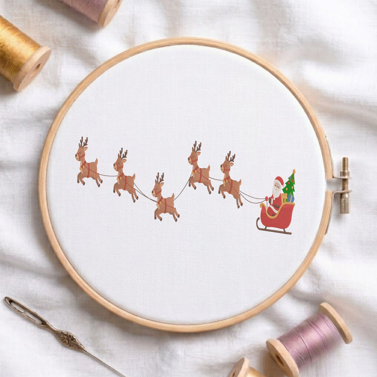 A circular embroidery hoop holding white fabric with a printed or stitched holiday scene showing Santa Claus in a red sleigh pulled by six brown reindeer, surrounded by spools of thread and a needle on a white fabric background.
