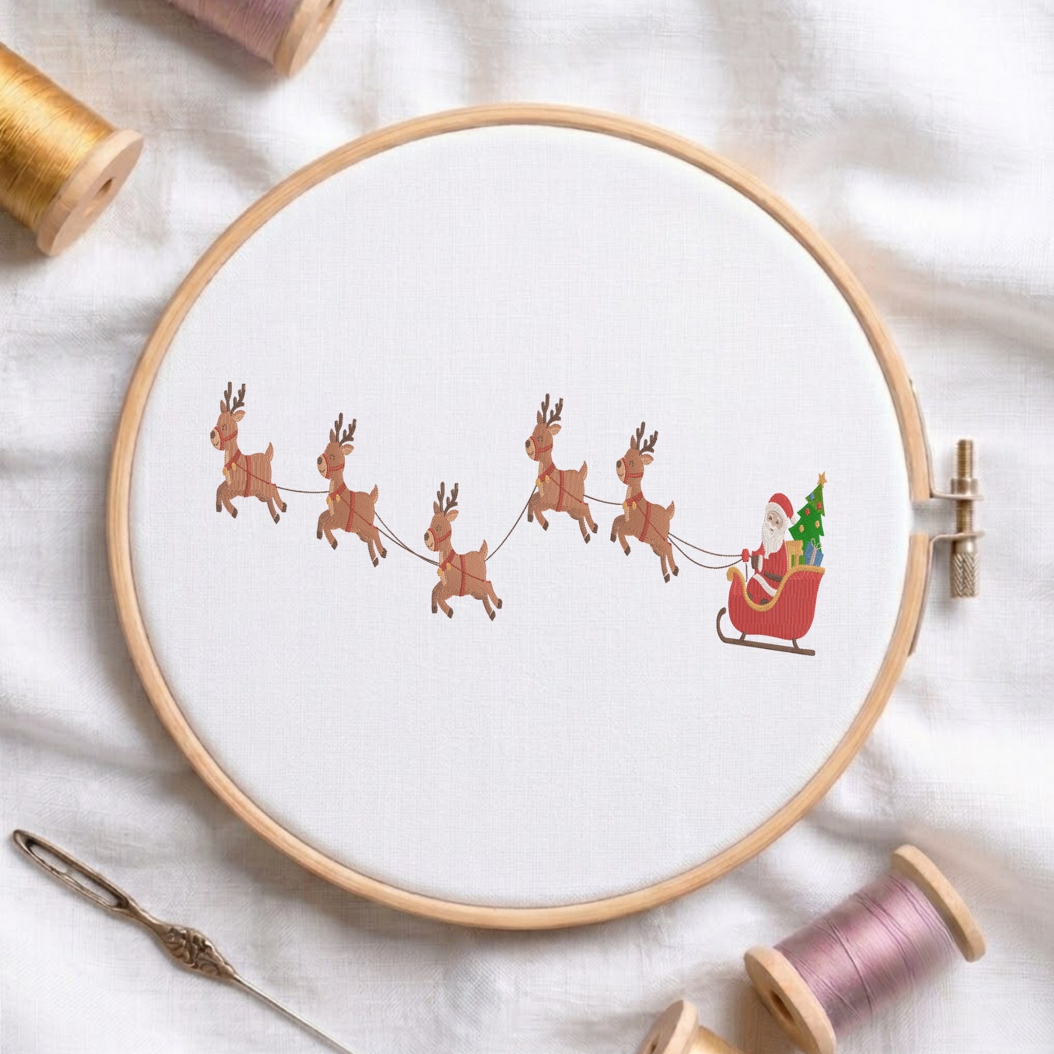 A circular embroidery hoop holding white fabric with a printed or stitched holiday scene showing Santa Claus in a red sleigh pulled by six brown reindeer, surrounded by spools of thread and a needle on a white fabric background.