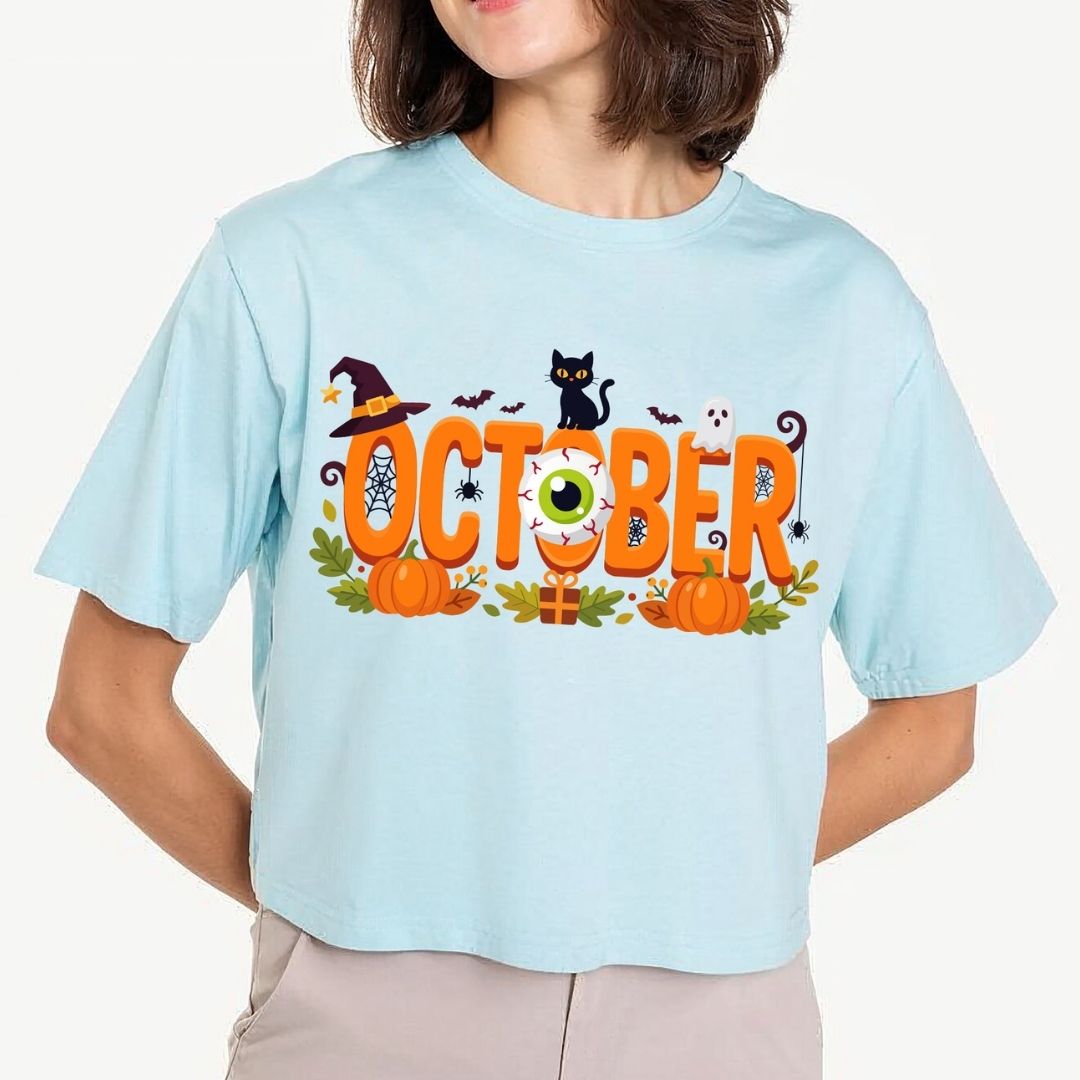 Woman wearing a light blue t-shirt featuring a spooky "October" typography design with a witch hat, black cat, and eyeball graphic.