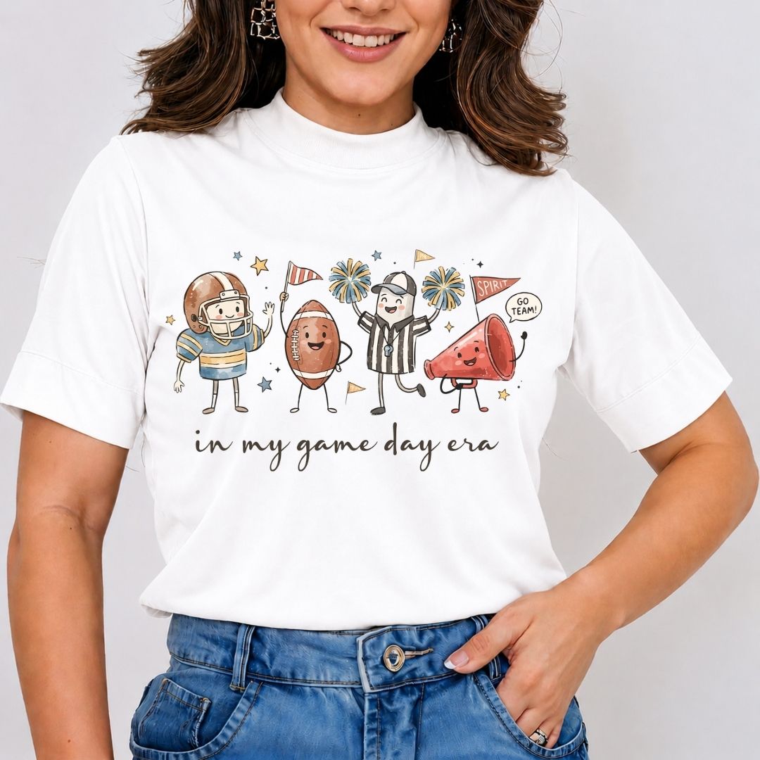 A woman wearing a white short-sleeve graphic t-shirt with a cartoon football-themed design and the text 'in my game day era', paired with blue jeans against a light gray background.