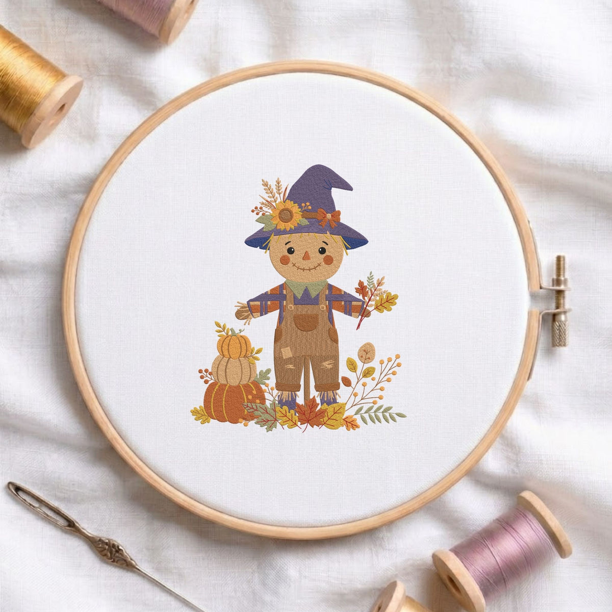 Cute autumn scarecrow wearing a witch hat with pumpkins and fall leaves machine embroidery design shown in a wooden embroidery hoop on white fabric with thread spools and a needle   
