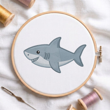 A completed embroidery design of a cartoon shark in a wooden hoop, surrounded by spools of thread and a needle on a white fabric background.