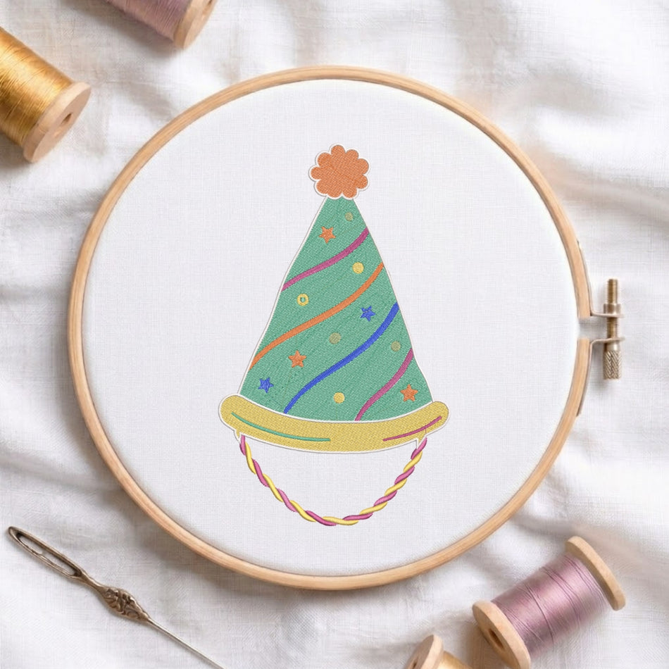 A circular embroidery hoop on white fabric, featuring a colorful embroidered party hat design. Surrounding props include spools of thread, a needle, and a metal eye pin on a soft white fabric background.