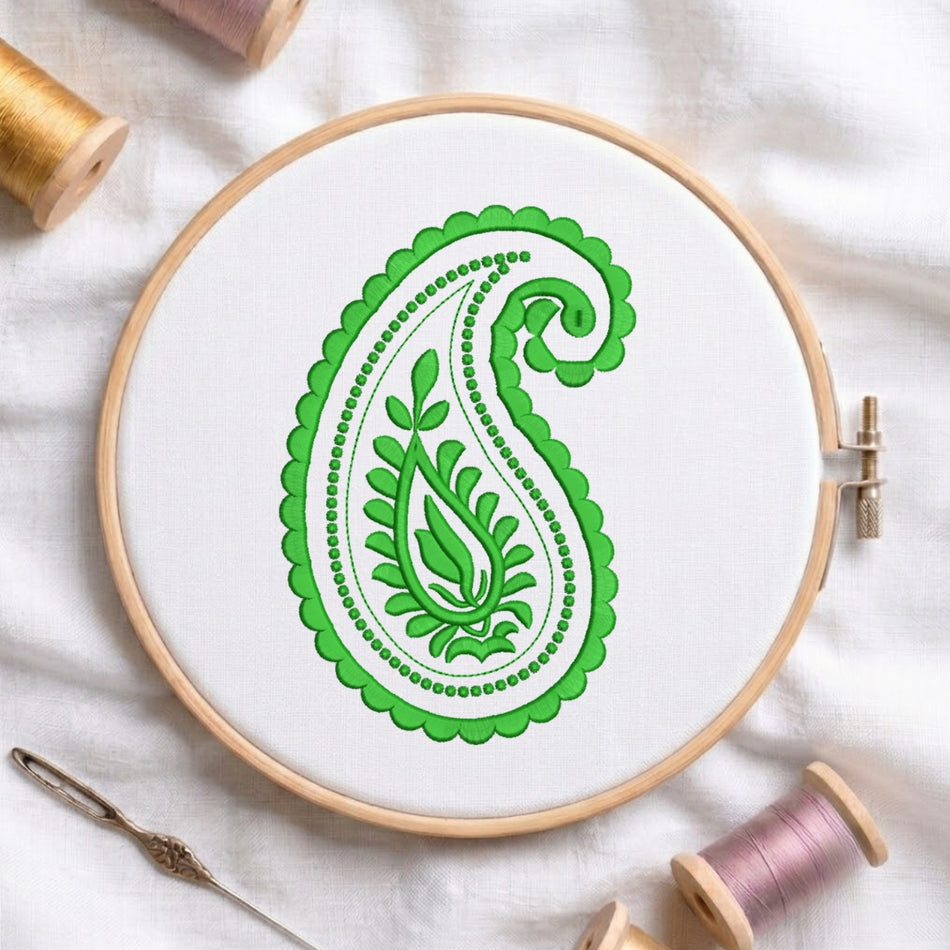 A circular embroidery hoop holding white fabric with a green paisley design, surrounded by spools of thread and a needle on a white fabric background.