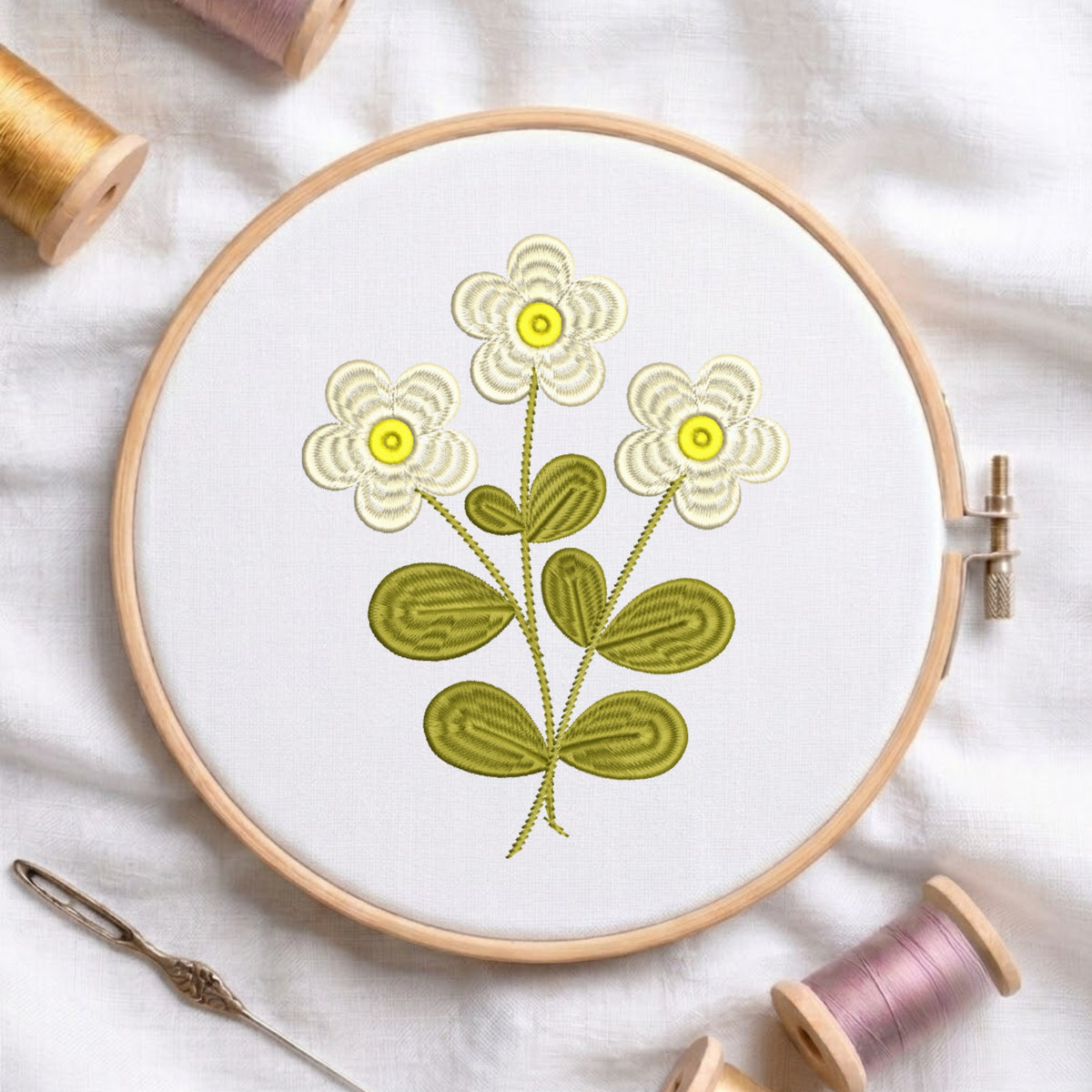 A round embroidery hoop holding white fabric with a stitched floral design of three cream flowers with yellow centers and olive green leaves on a white cloth background, surrounded by spools of thread and a needle.