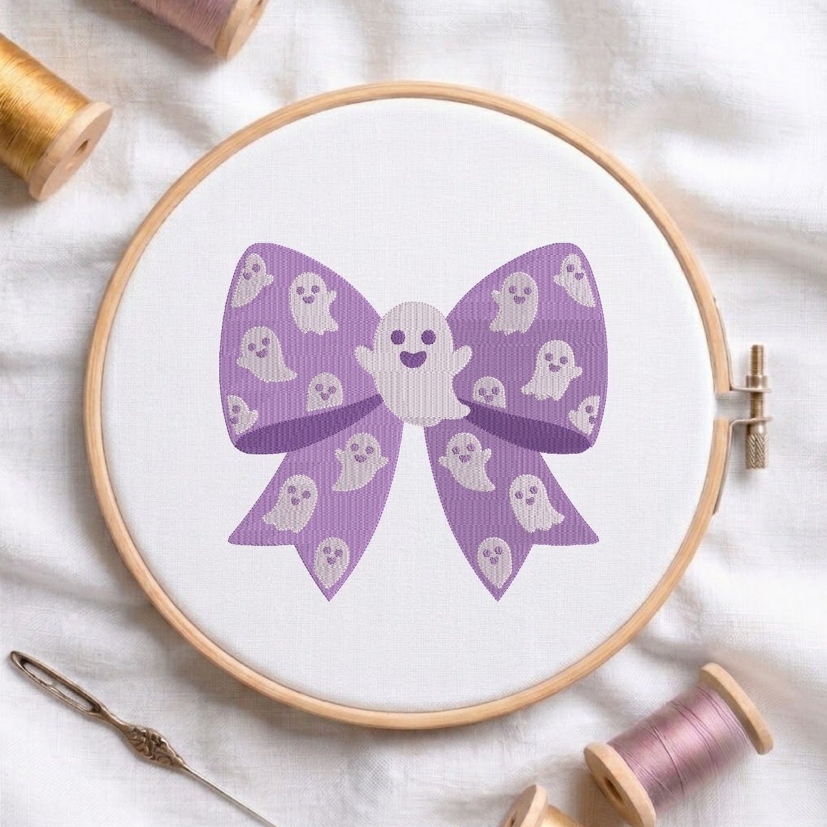 A white fabric stretched in a wooden embroidery hoop, featuring an embroidered design of a purple patterned bow filled with cute little ghost characters and a central larger ghost figure.