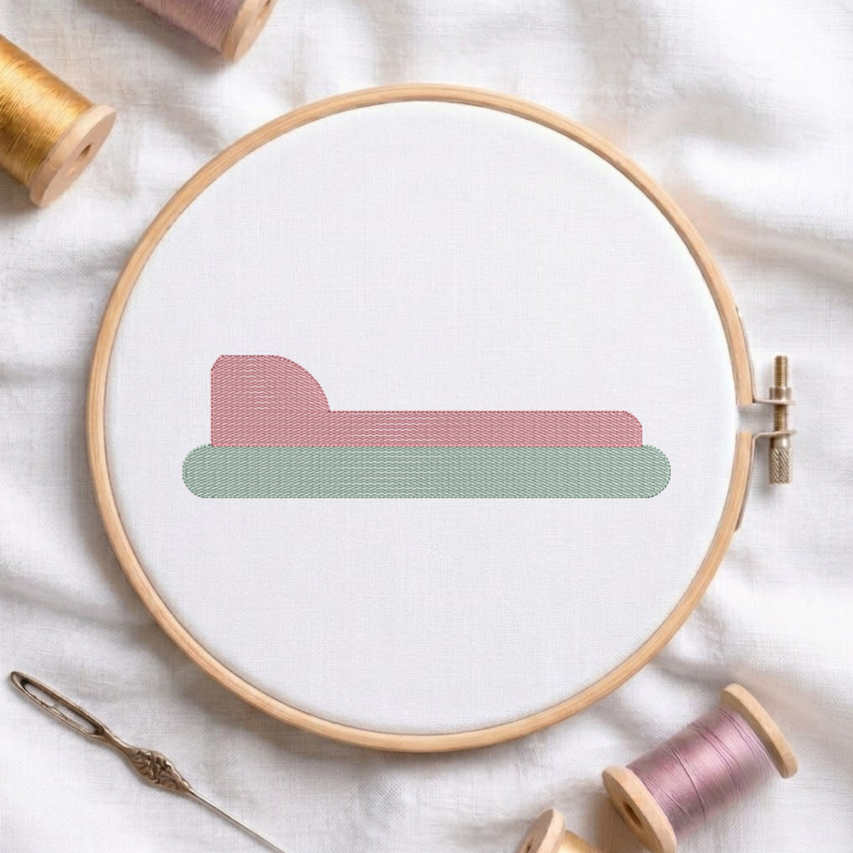 An embroidered minimalist bed design featuring a pink headboard and green base, stitched inside a wooden hoop on white fabric with sewing threads.
