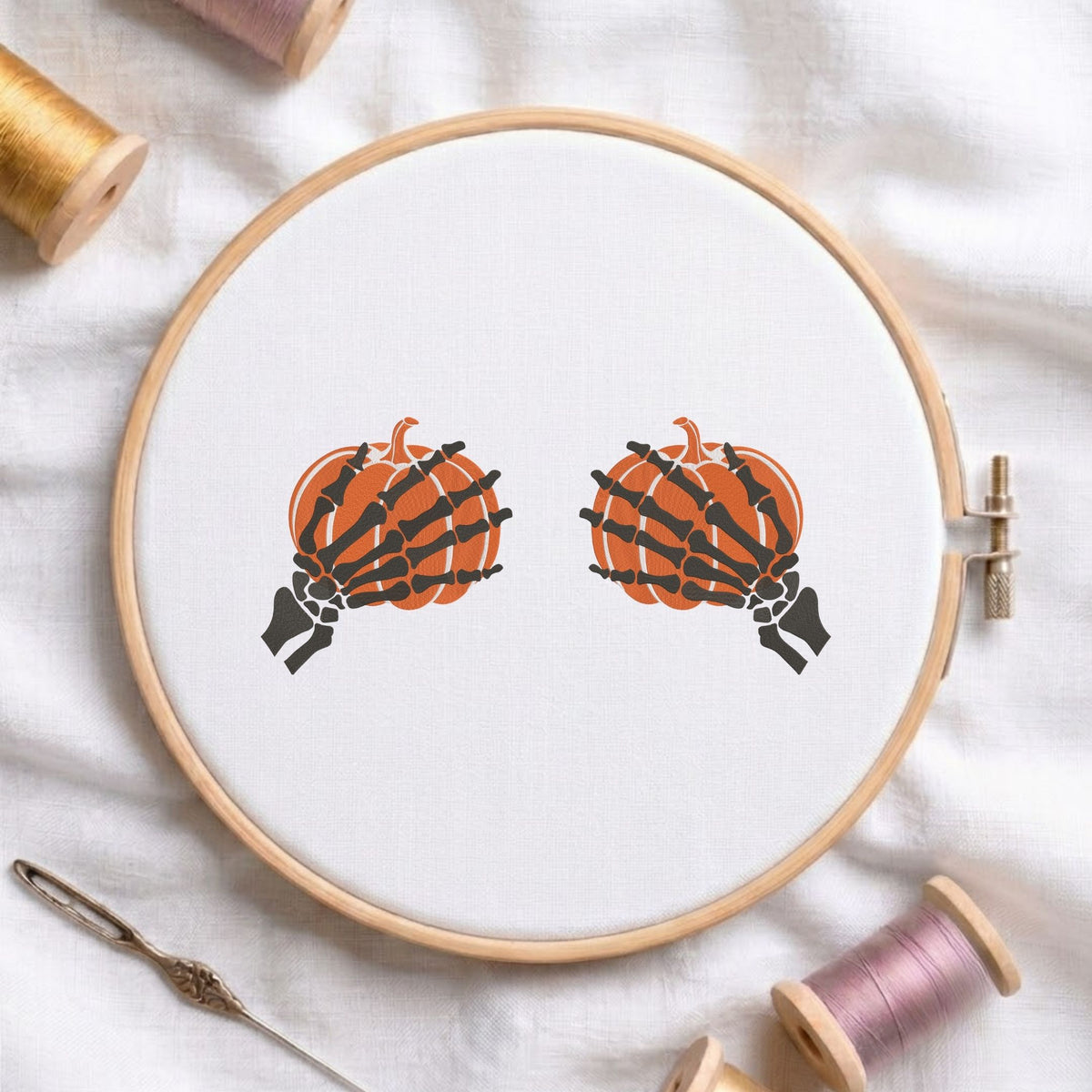 Skeleton hands holding pumpkins Halloween machine embroidery design shown in a wooden embroidery hoop on white fabric with thread spools and a needle   