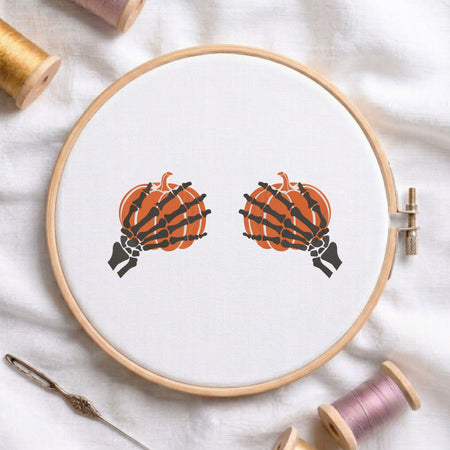 Skeleton hands holding pumpkins Halloween machine embroidery design shown in a wooden embroidery hoop on white fabric with thread spools and a needle   