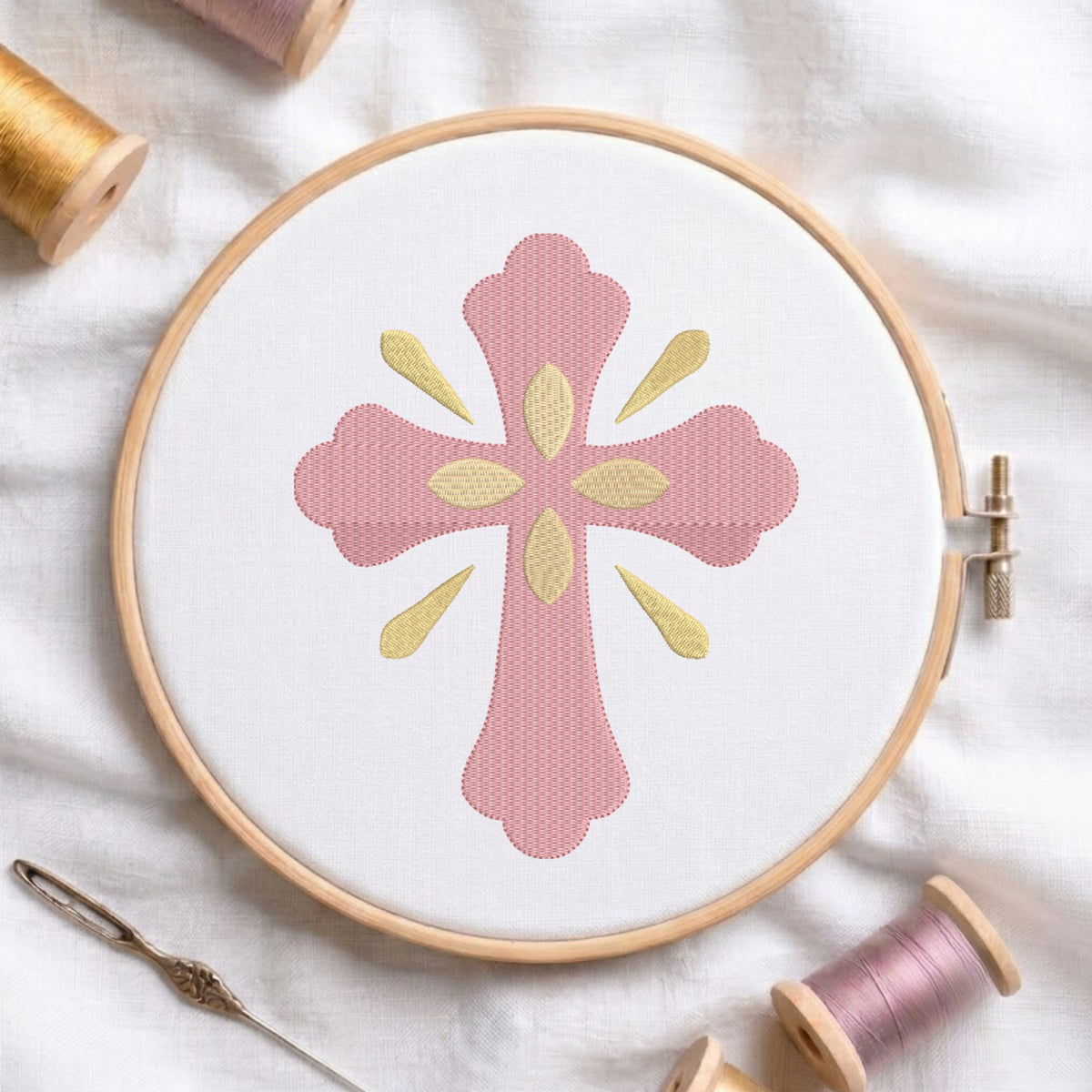 Soft pink ornamental cross with gold central petals and ray accents embroidered on white fabric inside a wooden hoop.