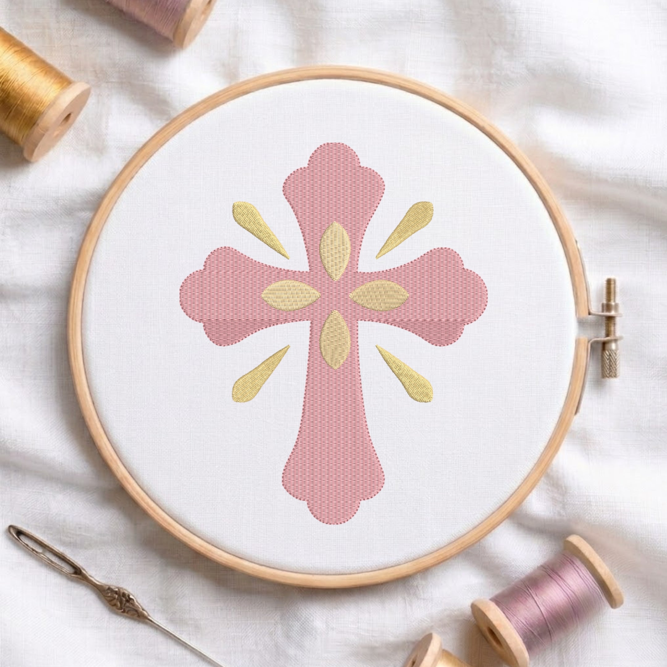 Soft pink ornamental cross with gold central petals and ray accents embroidered on white fabric inside a wooden hoop.