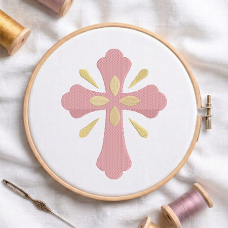 Soft pink ornamental cross with gold central petals and ray accents embroidered on white fabric inside a wooden hoop.