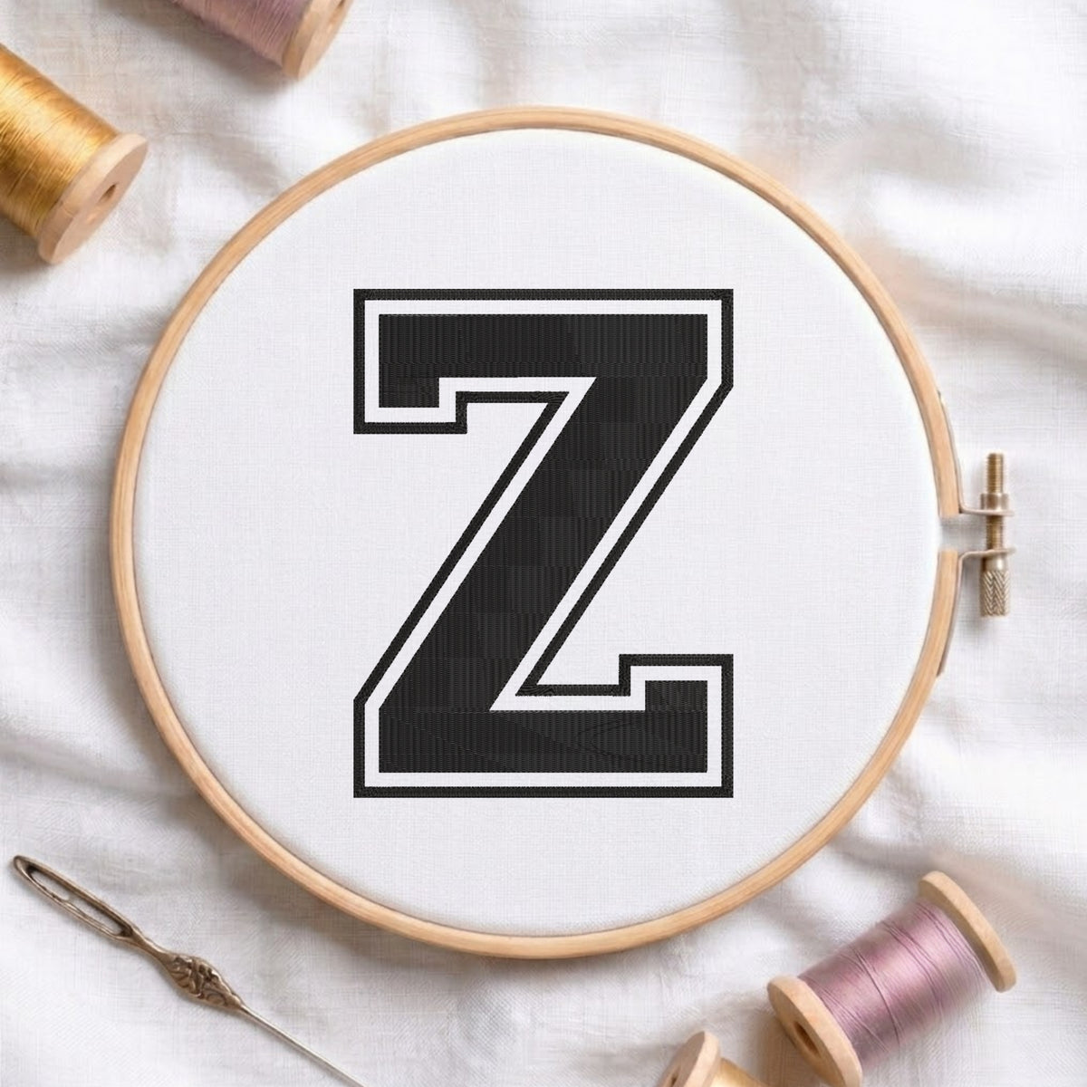 A wooden embroidery hoop holding white fabric with a large black letter Z outlined in white, styled on a white fabric surface with spools of thread and a needle.