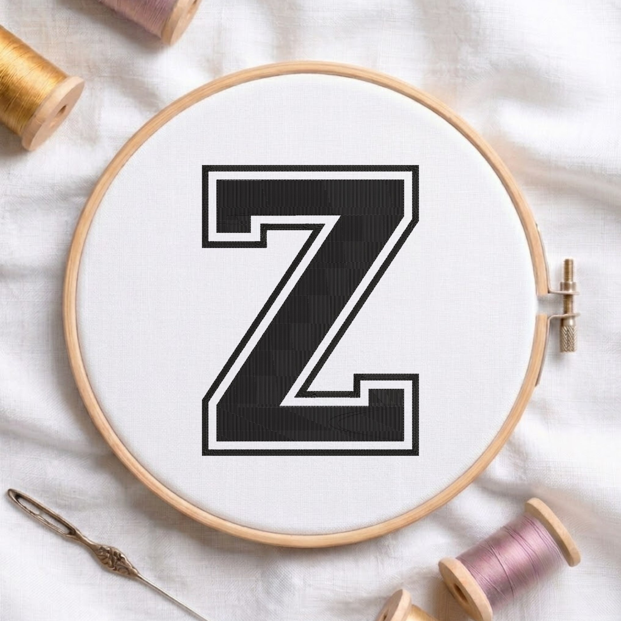 A wooden embroidery hoop holding white fabric with a large black letter Z outlined in white, styled on a white fabric surface with spools of thread and a needle.