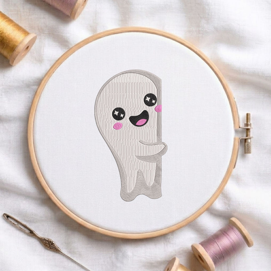 Cute grey ghost with a happy smiling face, pink cheeks, and star eyes machine embroidery design, displayed inside a round wooden hoop on white fabric with thread spools and a needle.
