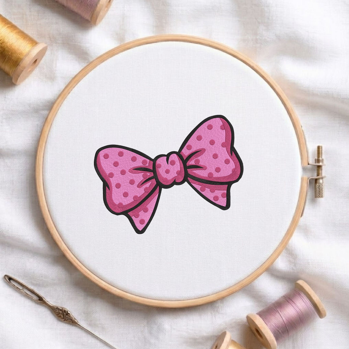 Pink polka dot bow machine embroidery design displayed in a wooden embroidery hoop on white fabric with thread spools and a needle
