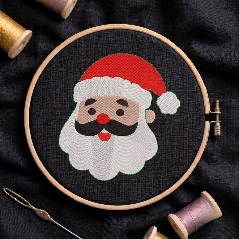 A circular embroidery hoop holding black fabric with a stitched Santa face design, surrounded by spools of thread and a needle on a dark fabric background.
