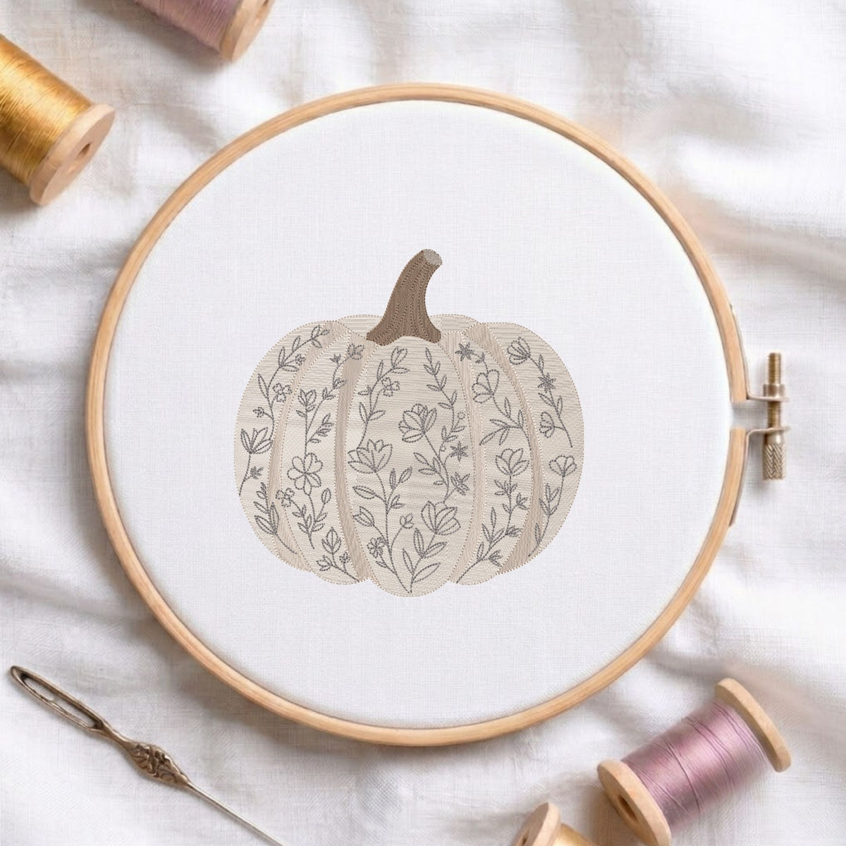 Floral patterned pumpkin machine embroidery design displayed in a wooden hoop on white fabric with thread spools and a needle.