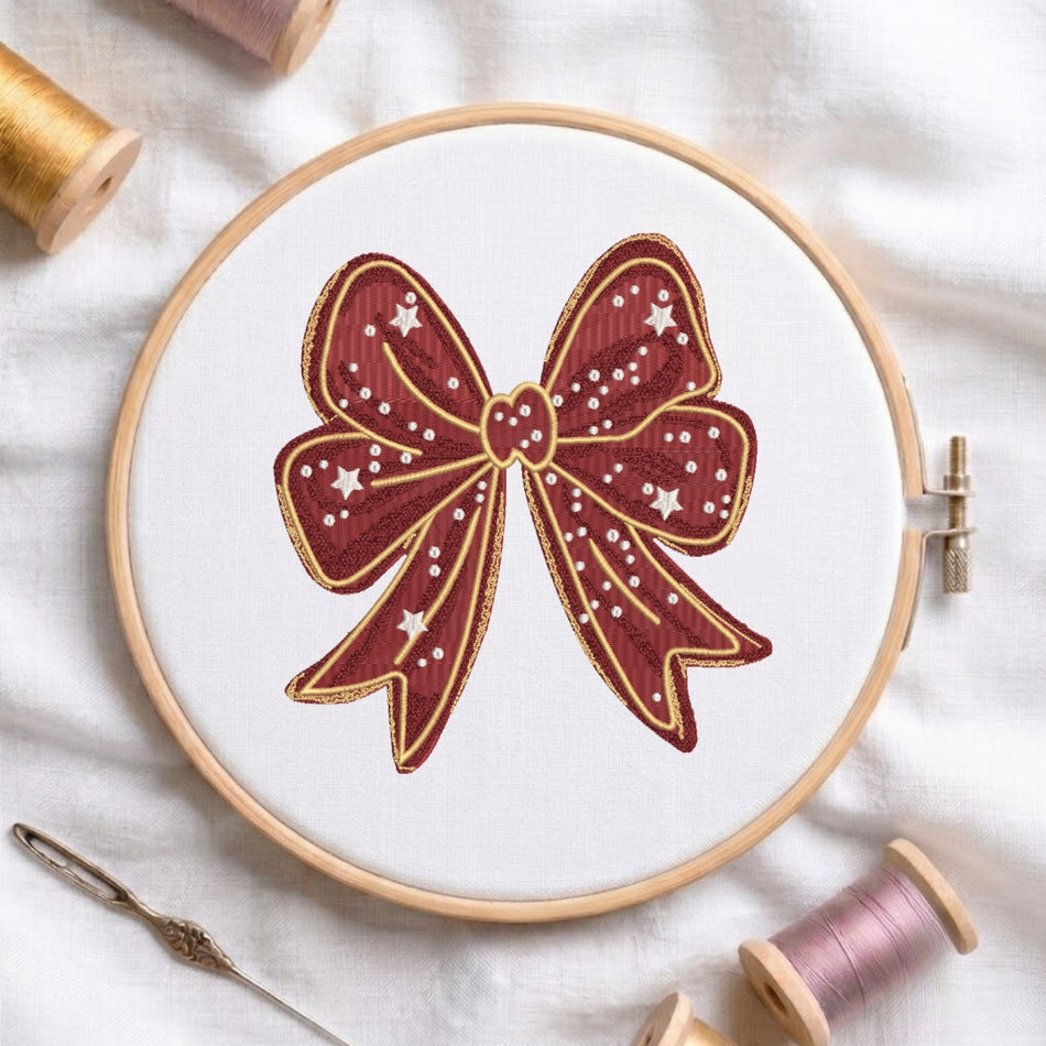 A circular embroidery hoop holding white fabric with a stitched bow design in deep red, gold, and white. The hoop is photographed on a white fabric surface with spools of thread and a needle around it.
