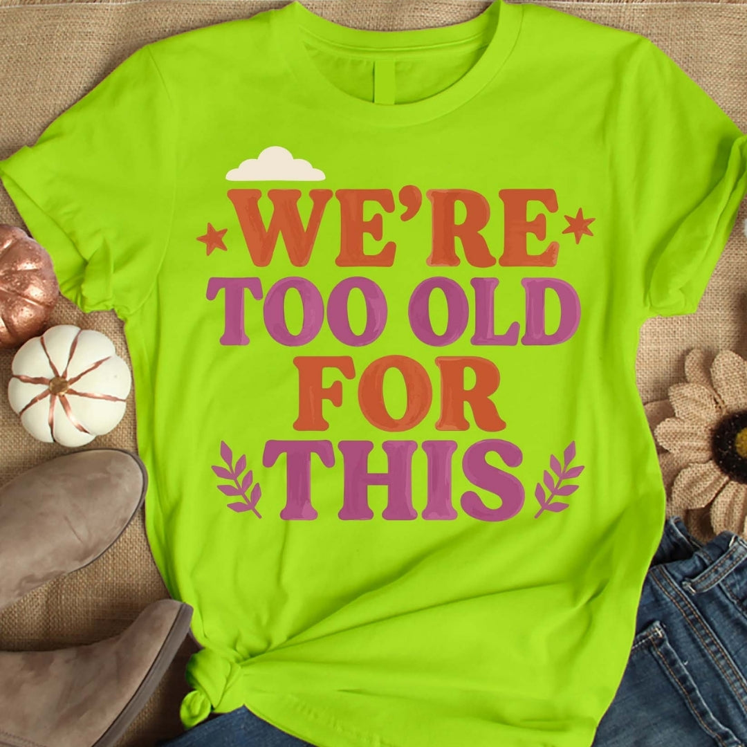 Lime green t-shirt with 'WE'RE TOO OLD FOR THIS' text on a textured surface.