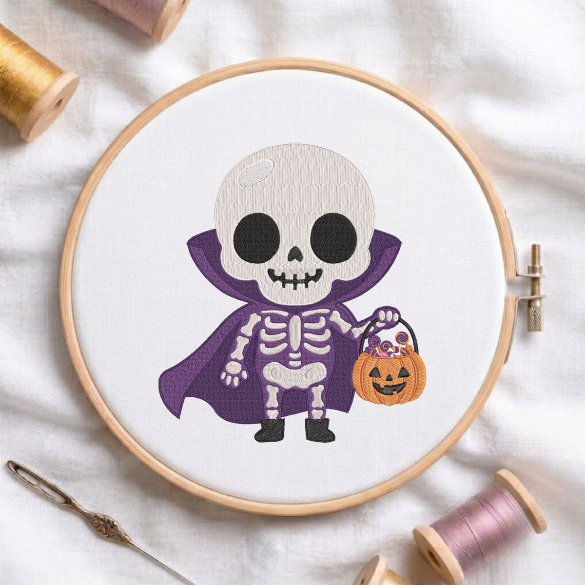 A circular embroidery hoop displays a cartoon skeleton dressed as a vampire holding a jack-o'-lantern bucket. The hoop is styled on a white fabric background with spools of thread and a needle.