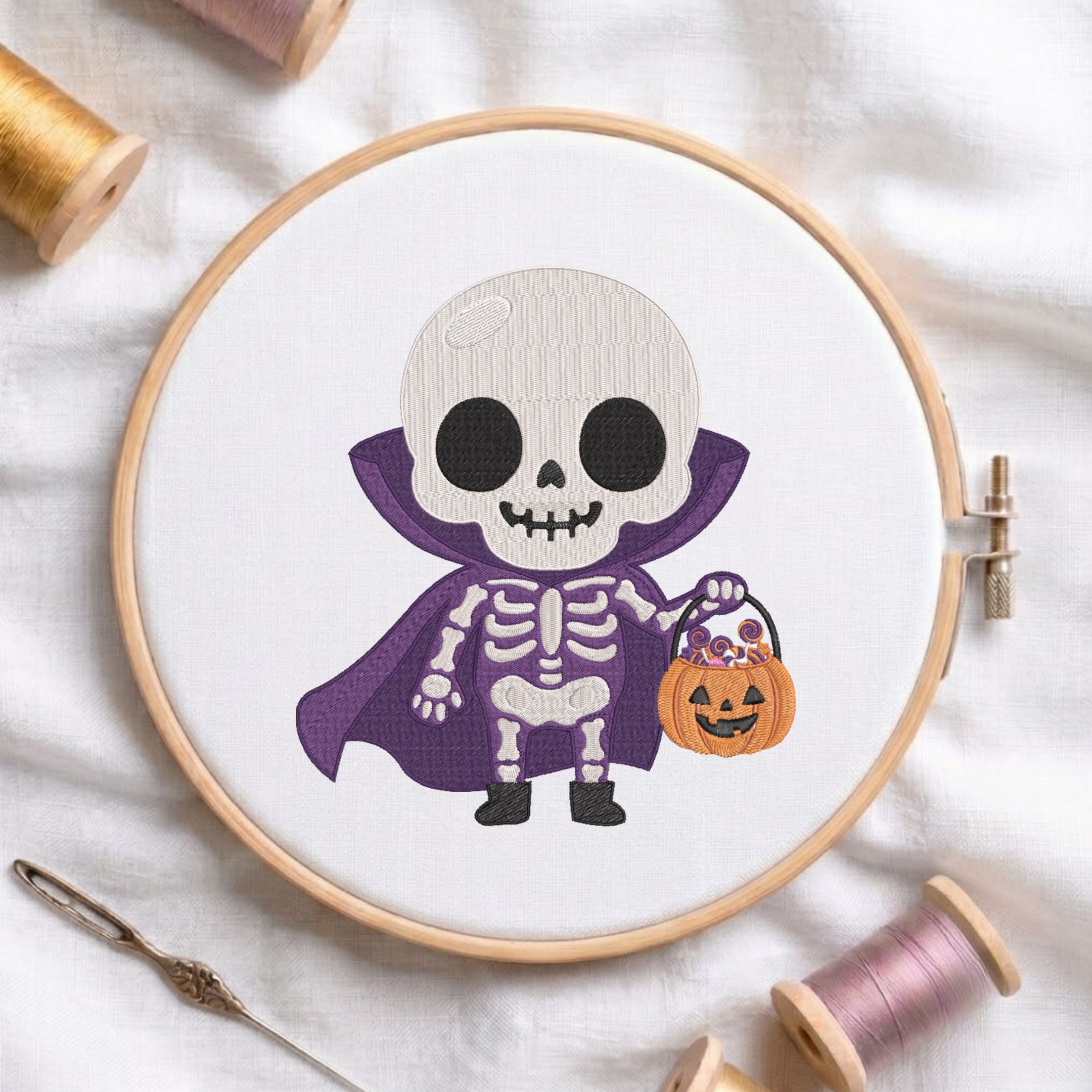 A circular embroidery hoop displays a cartoon skeleton dressed as a vampire holding a jack-o'-lantern bucket. The hoop is styled on a white fabric background with spools of thread and a needle.