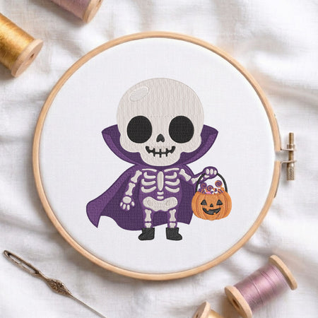 A circular embroidery hoop displays a cartoon skeleton dressed as a vampire holding a jack-o'-lantern bucket. The hoop is styled on a white fabric background with spools of thread and a needle.
